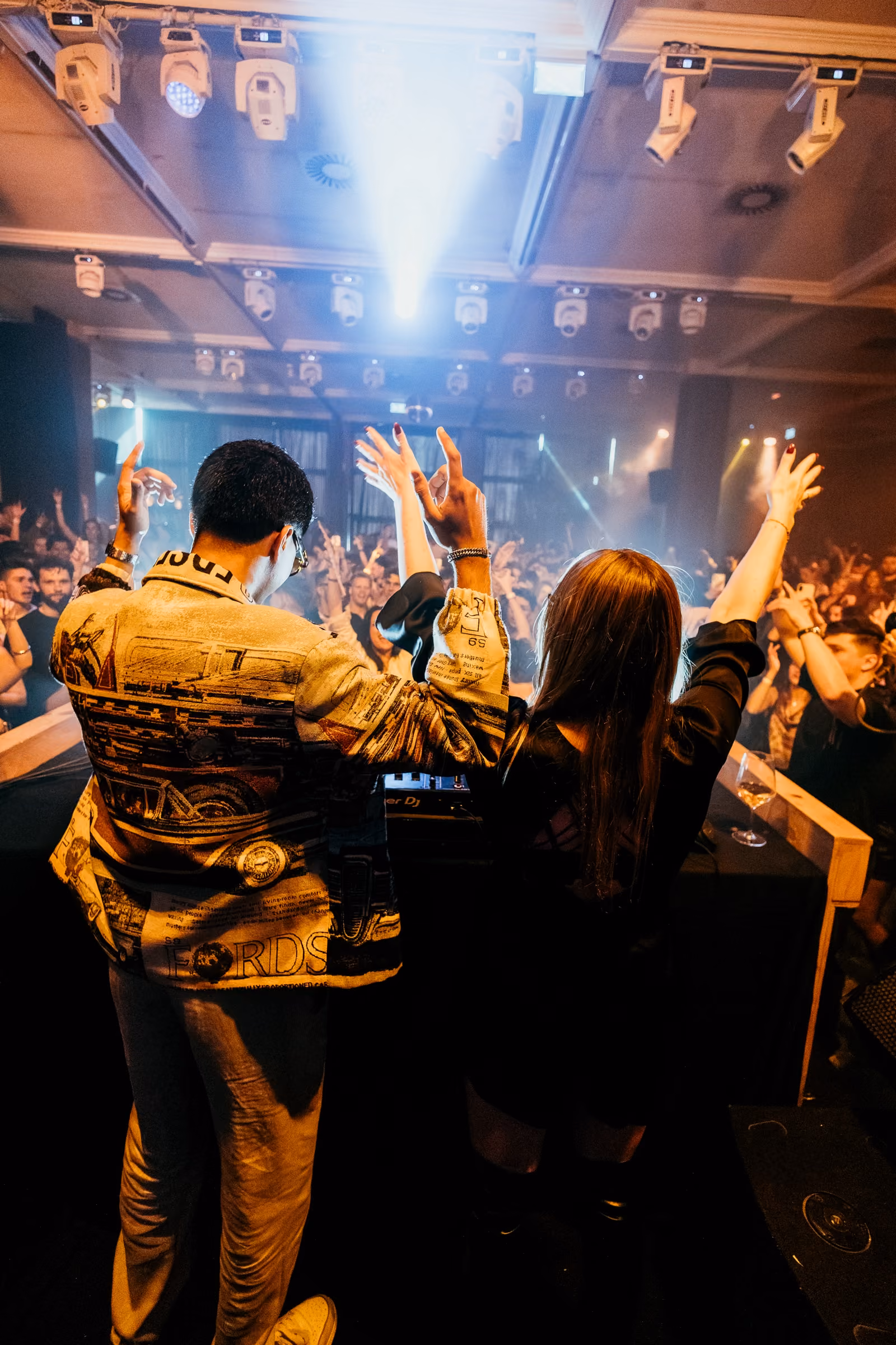 Yassine and Lisa behind the decks under gold light, the crowd in front of them