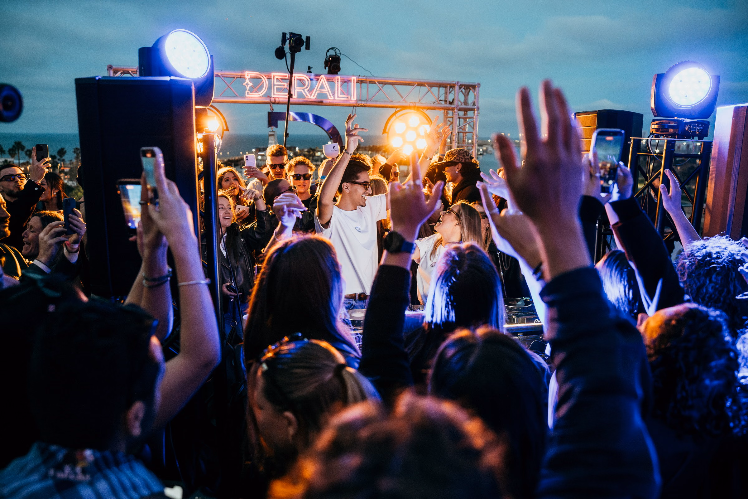 The Derali truss sign on the rooftop at dusk, hands and phones raised beneath it