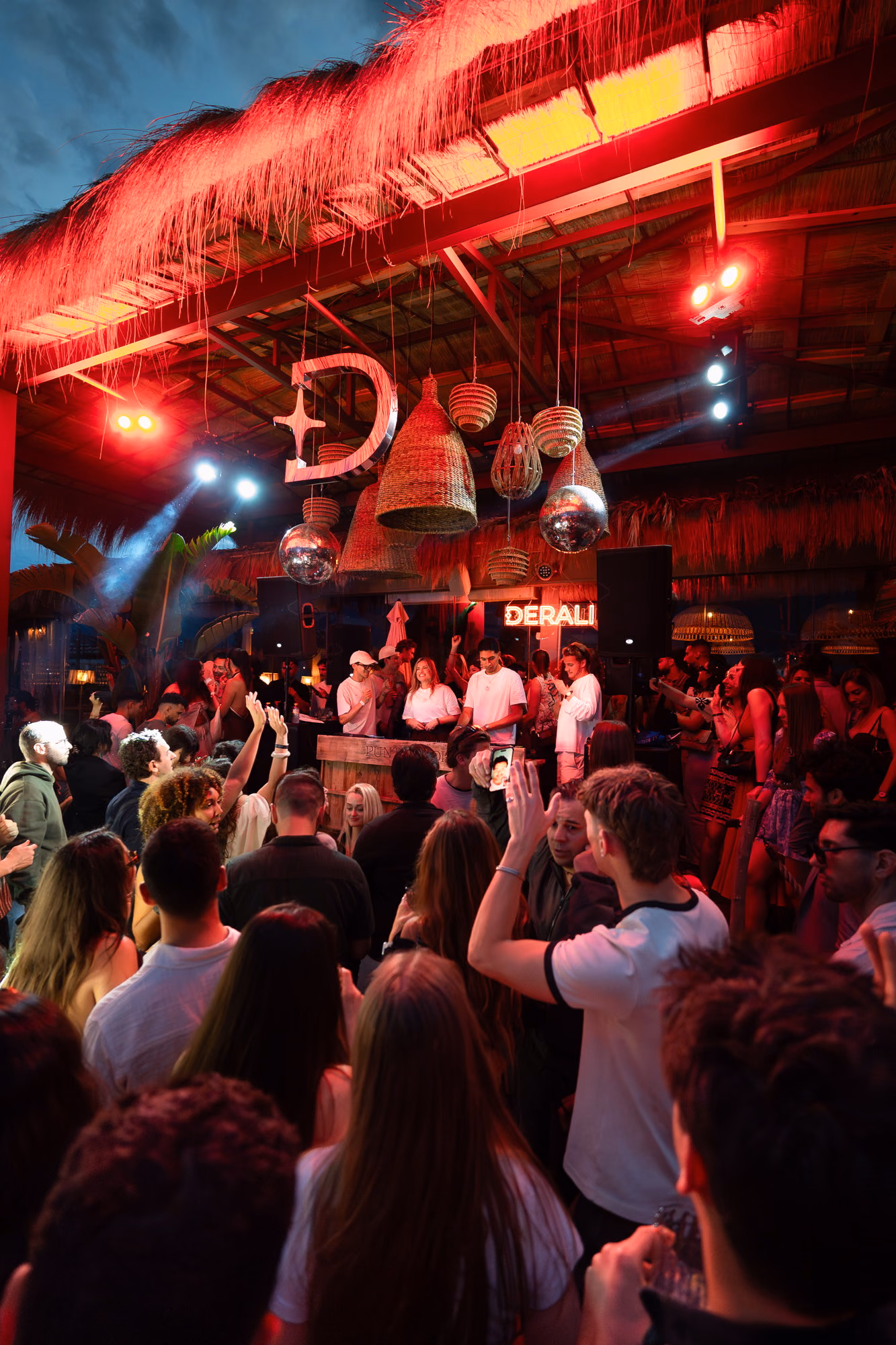 The red-lit Derali sign under a thatched beach-club roof, rattan lamps above the crowd