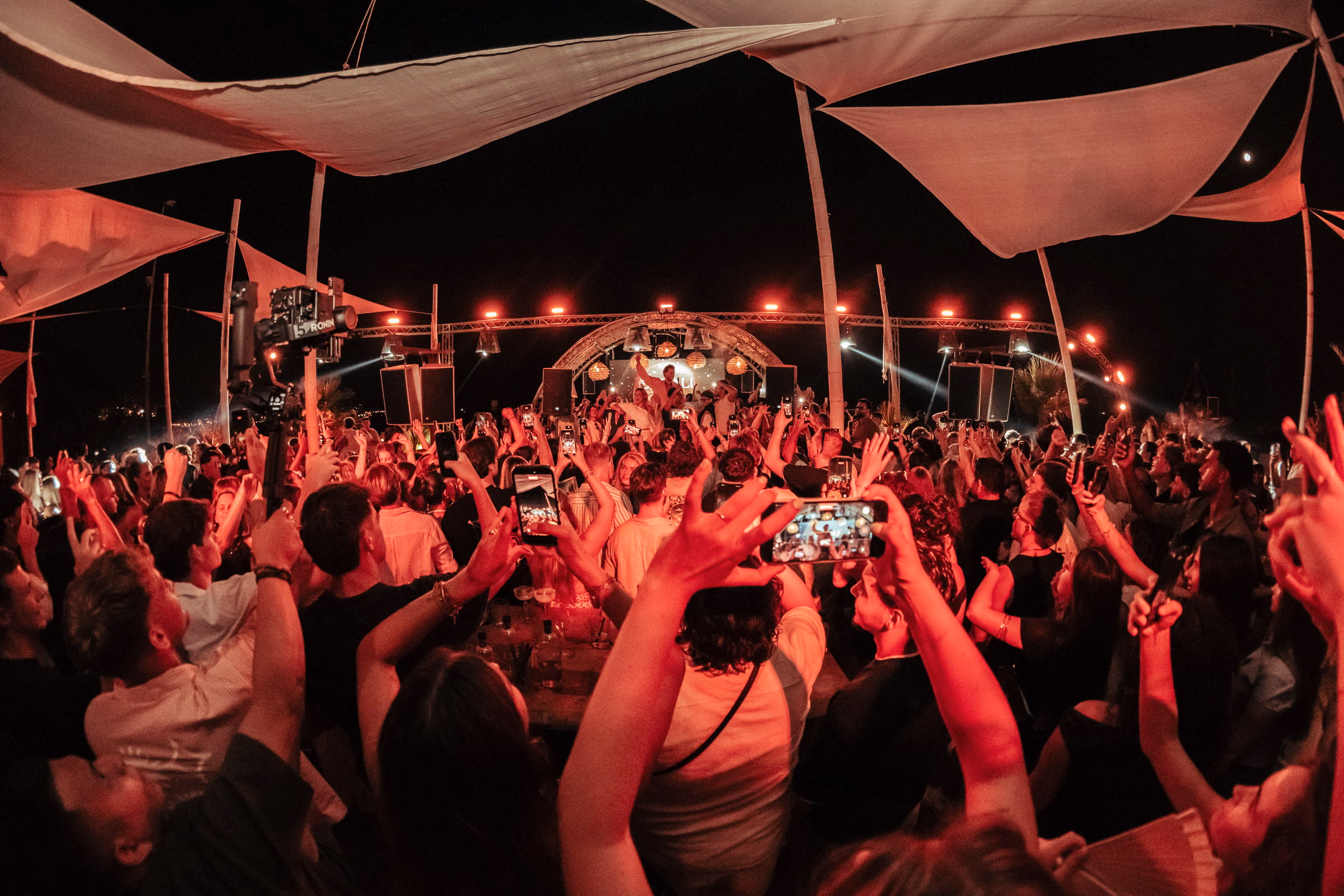 White sail canopies over a packed beach club, the whole floor lit red