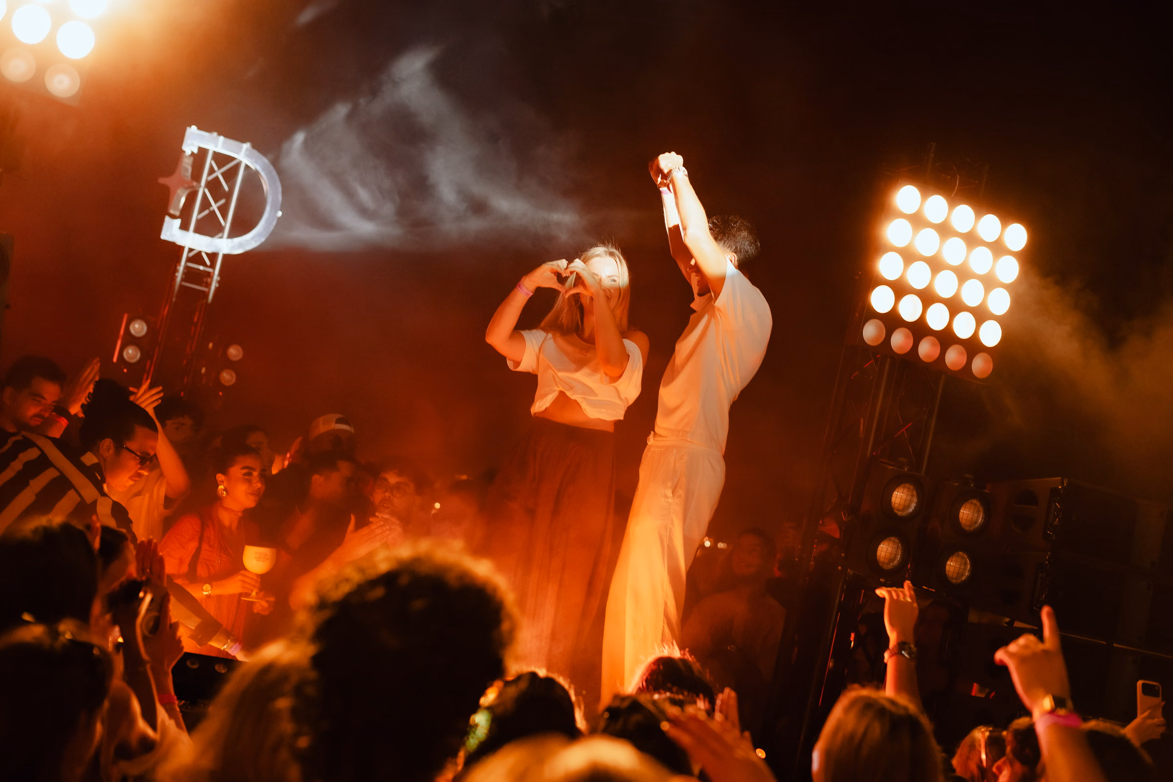 A jet of smoke crossing the open-air stage, two figures in white at the booth