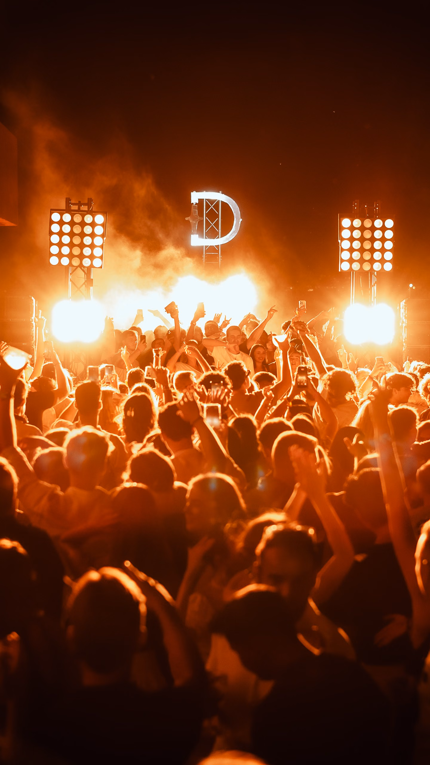 An open-air crowd in front of the lit Derali monogram, flame and orange haze behind the booth