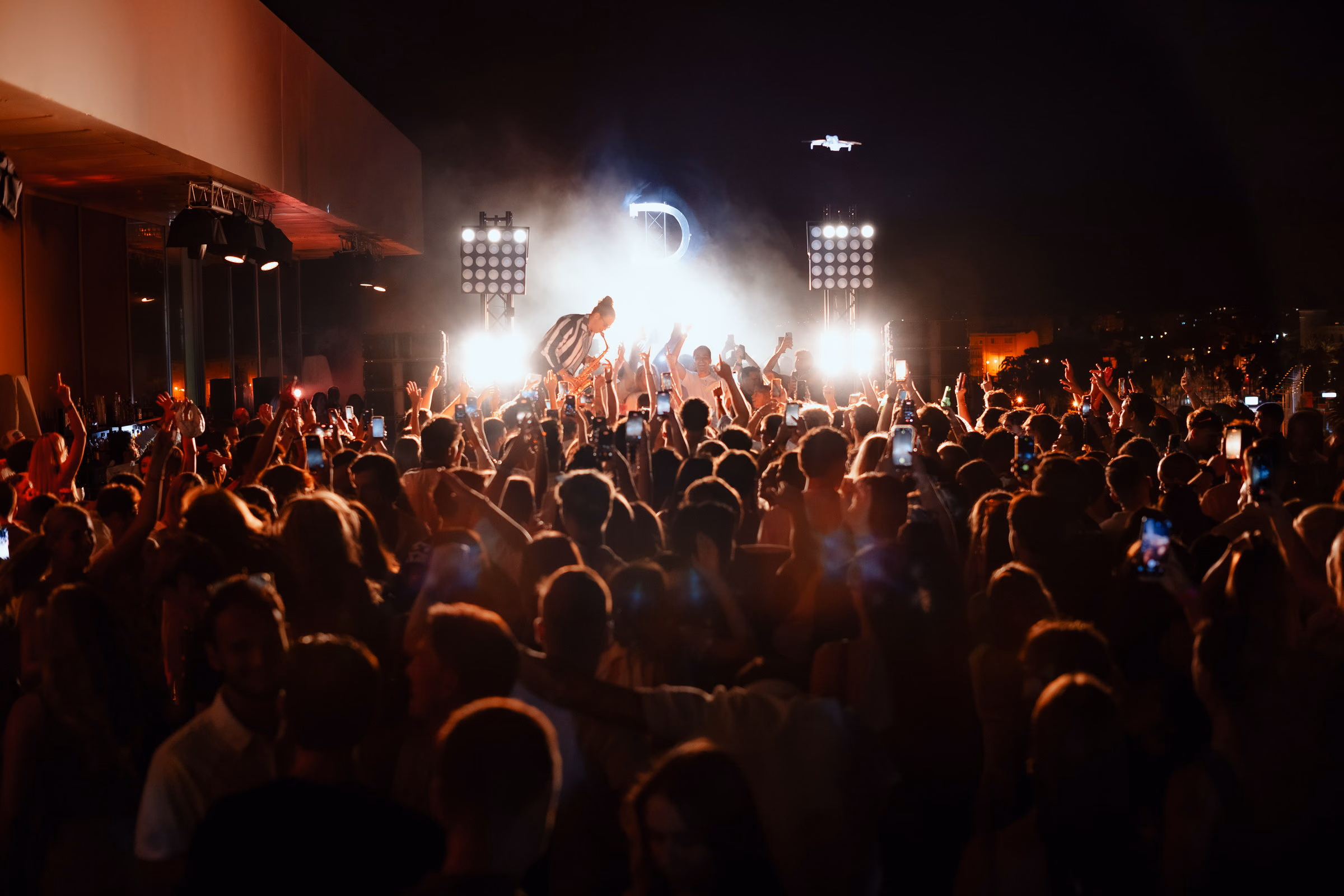 An open-air crowd at night, blinders and smoke around the lit Derali monogram