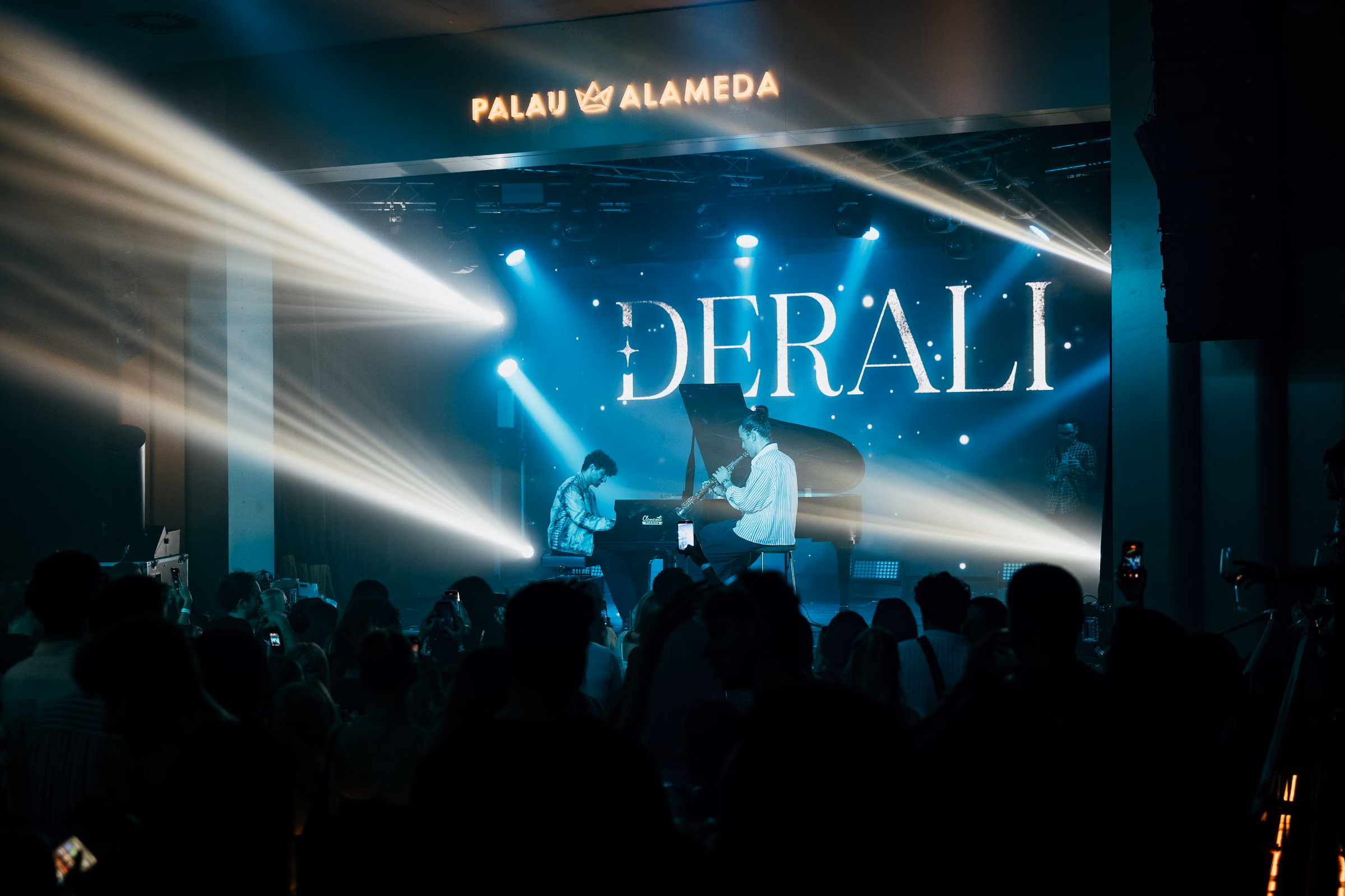 A pianist at the grand piano beneath the Derali wordmark at Palau Alameda, blue beams crossing