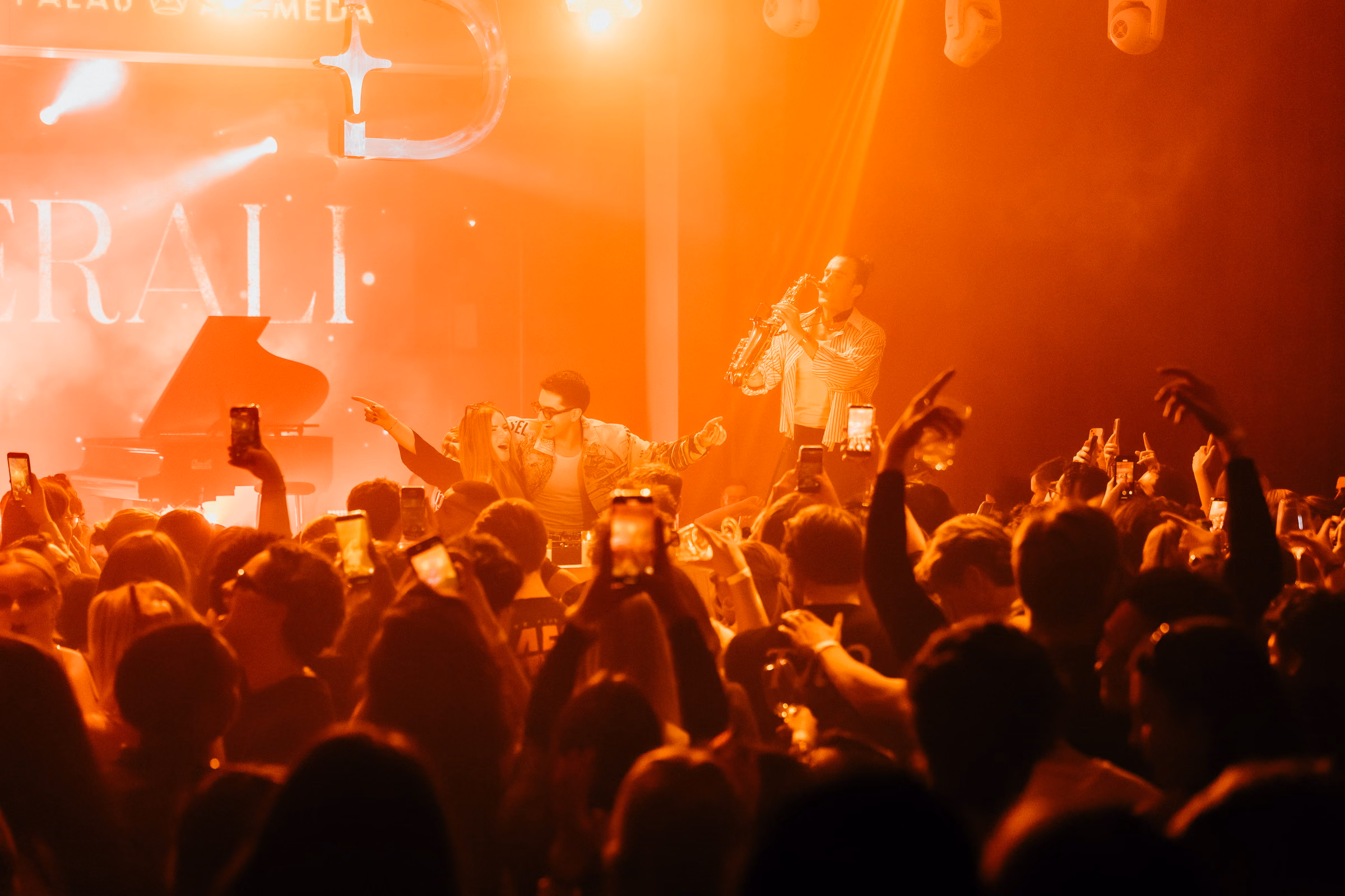 A saxophonist on stage above a packed floor at Palau Alameda, lit amber