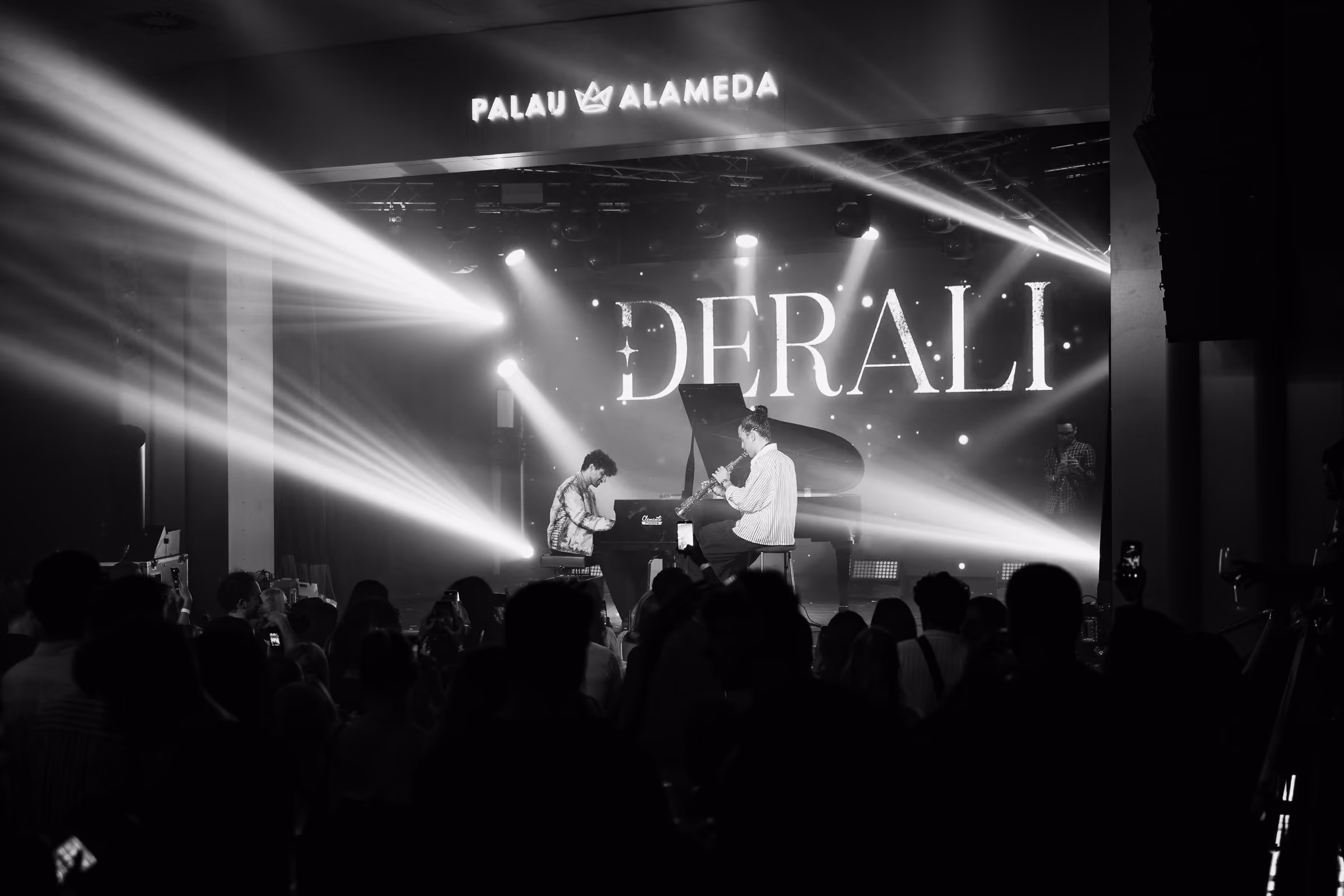 A saxophonist and a pianist at the grand piano under the Derali wordmark, in black and white
