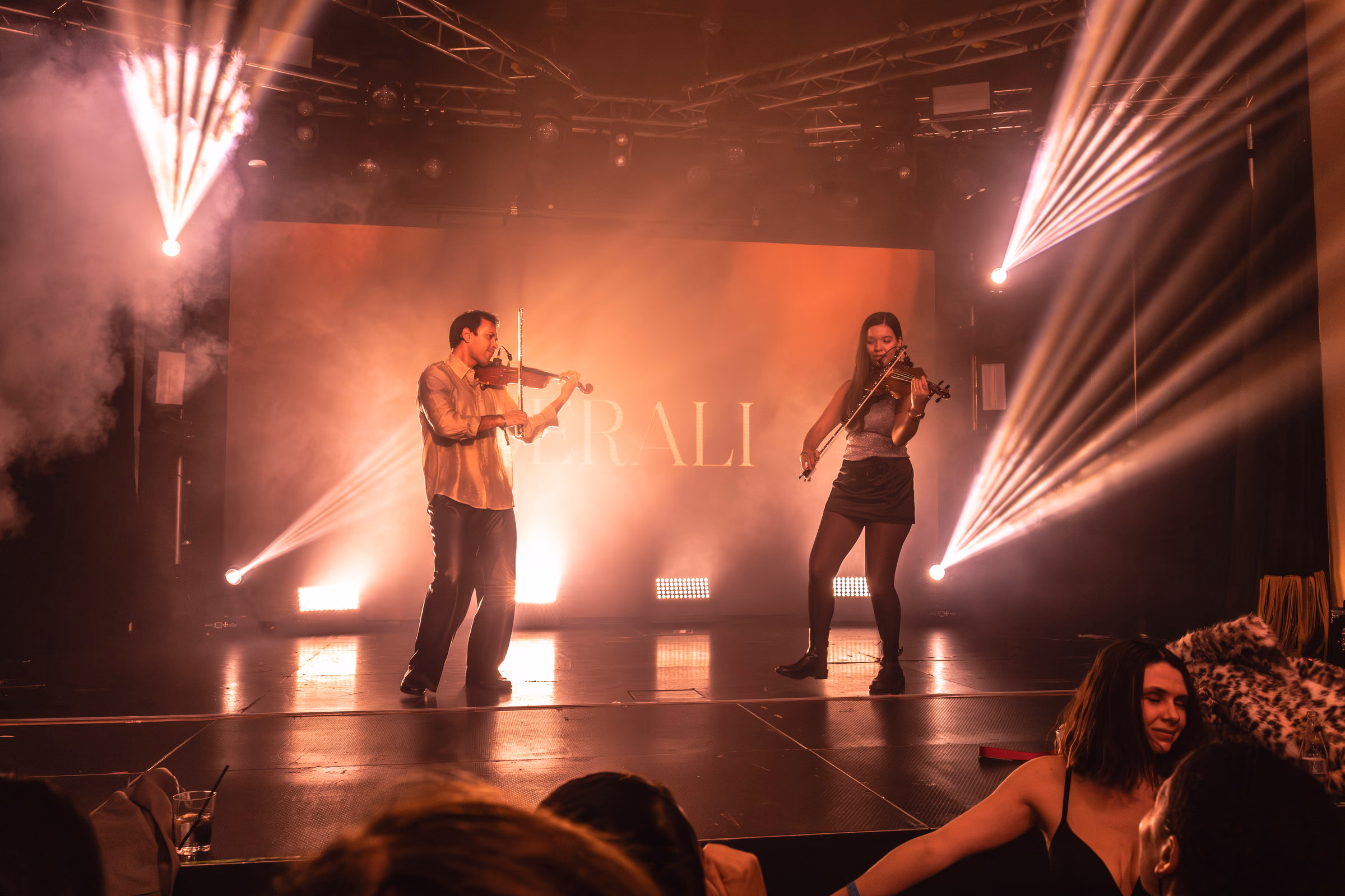 Two violinists playing either side of the Derali wordmark, amber beams behind them