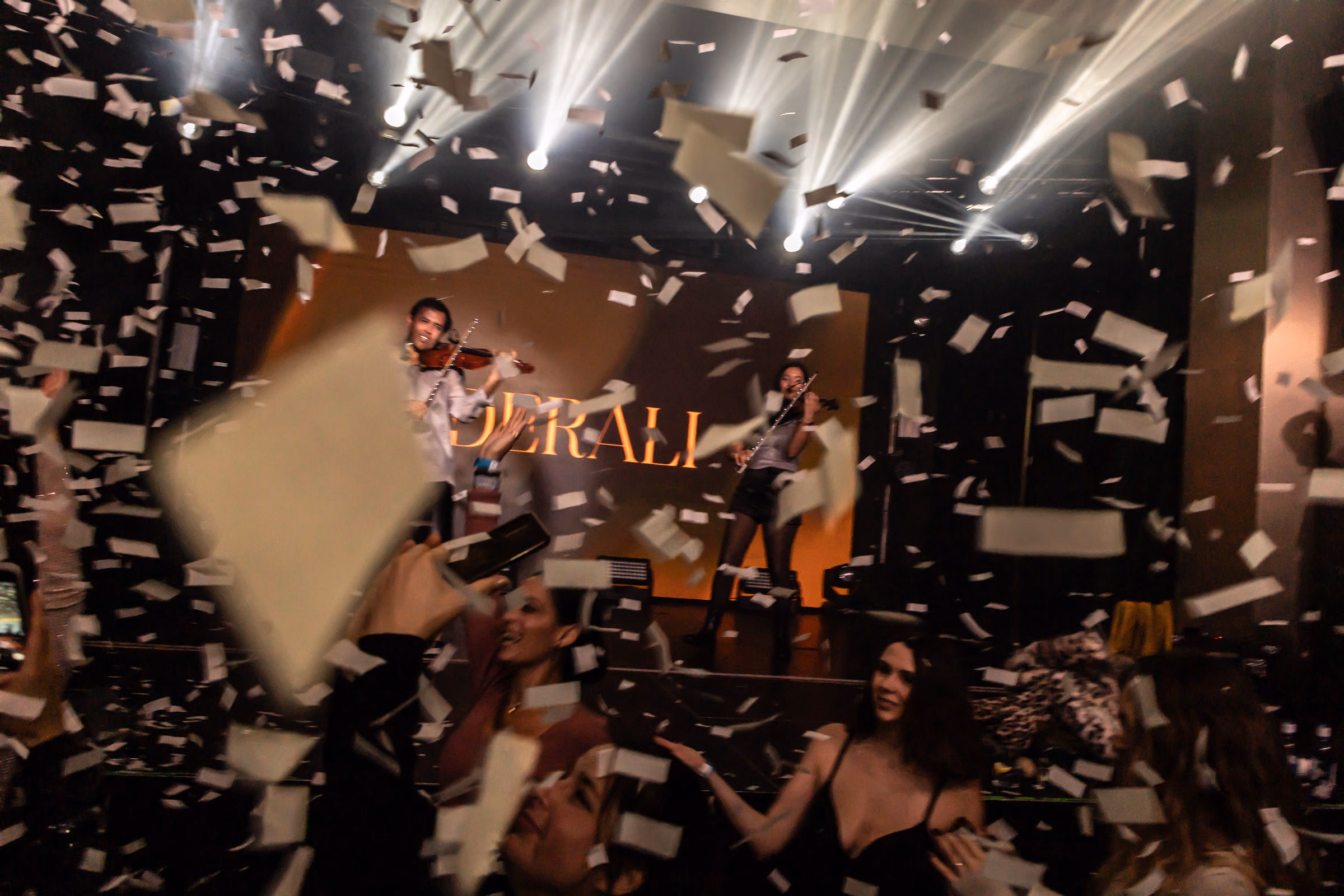 Two violinists playing through a fall of white confetti, the crowd filming