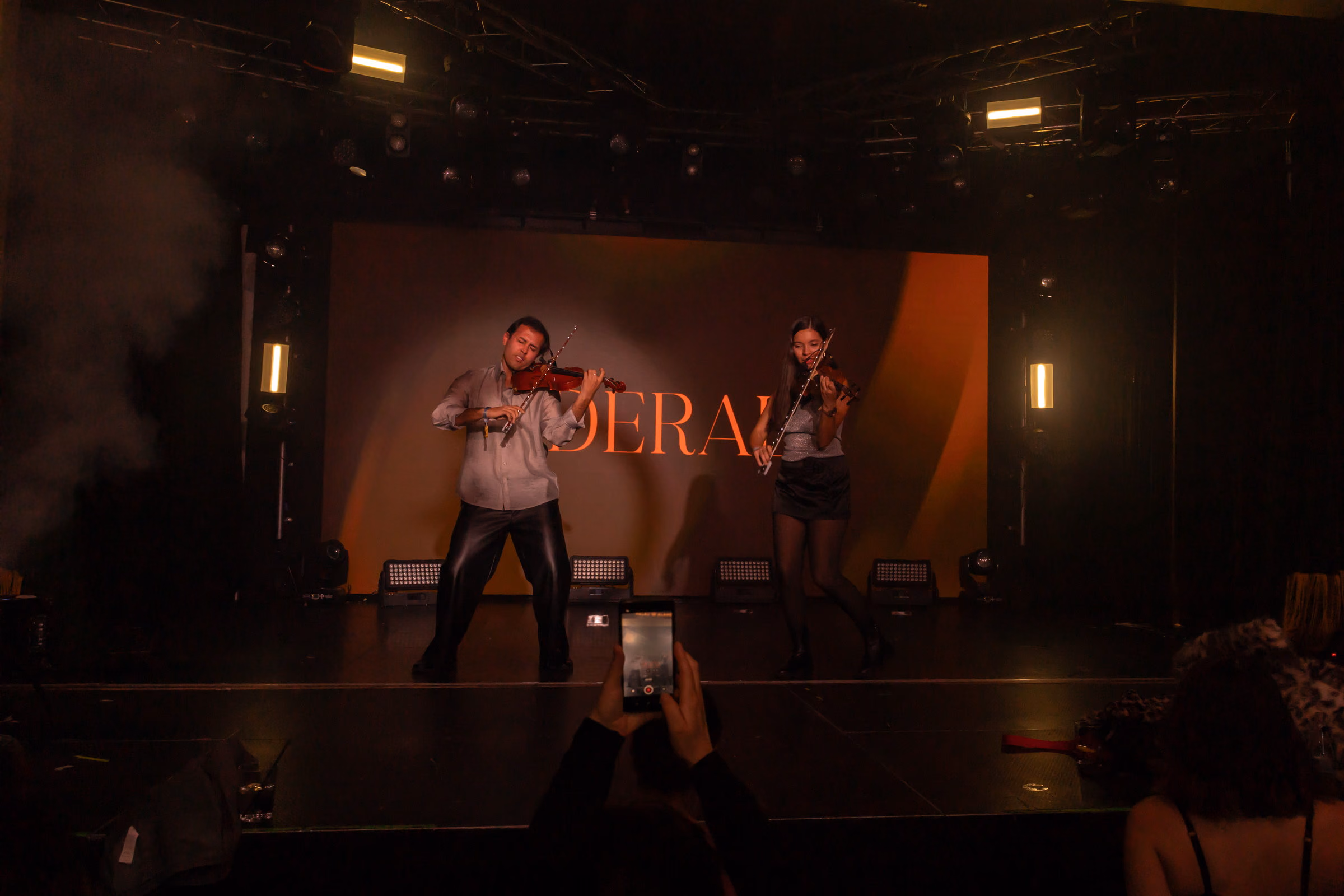 Two violinists apart on a wide stage in front of the Derali wordmark